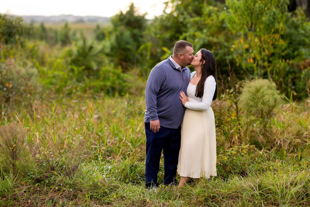 A couple embraces in a scenic field with tall grass and wildflowers during a romantic outdoor photo session.