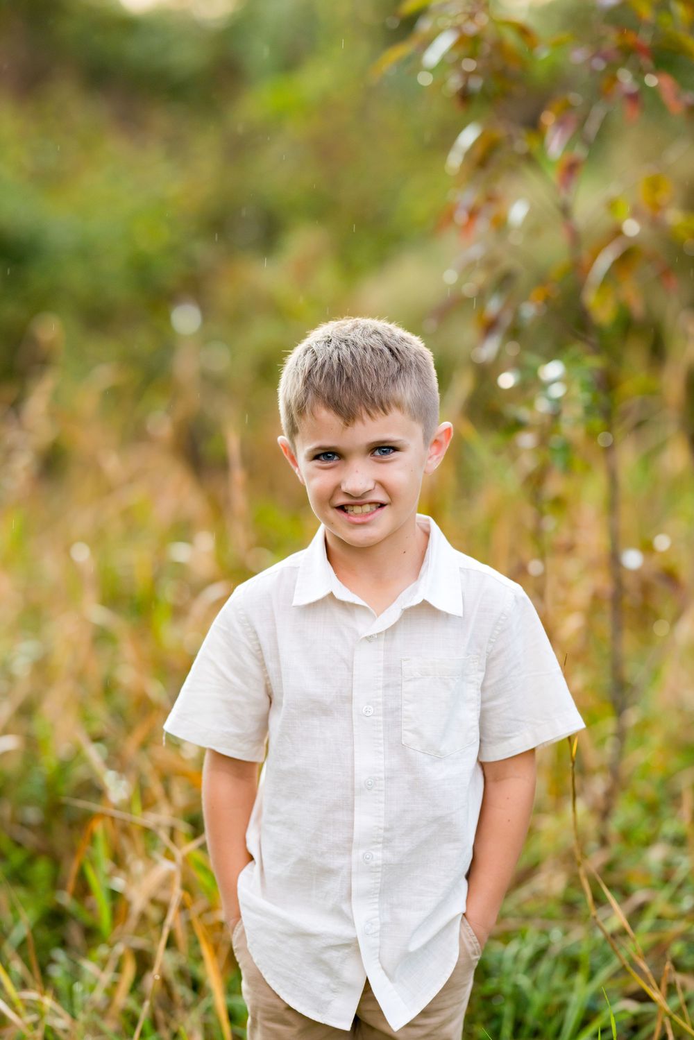 Child in white button-down shirt poses outdoors against blurred natural background with autumn foliage.