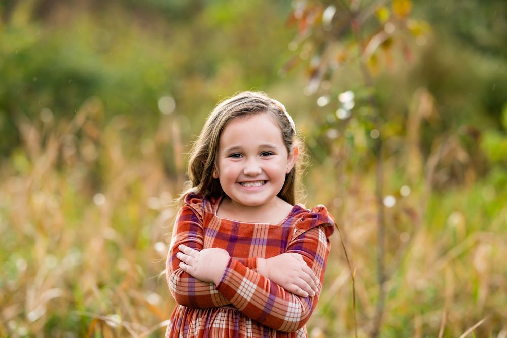 A person in a red plaid dress poses outdoors in a natural setting with autumn foliage in the background.