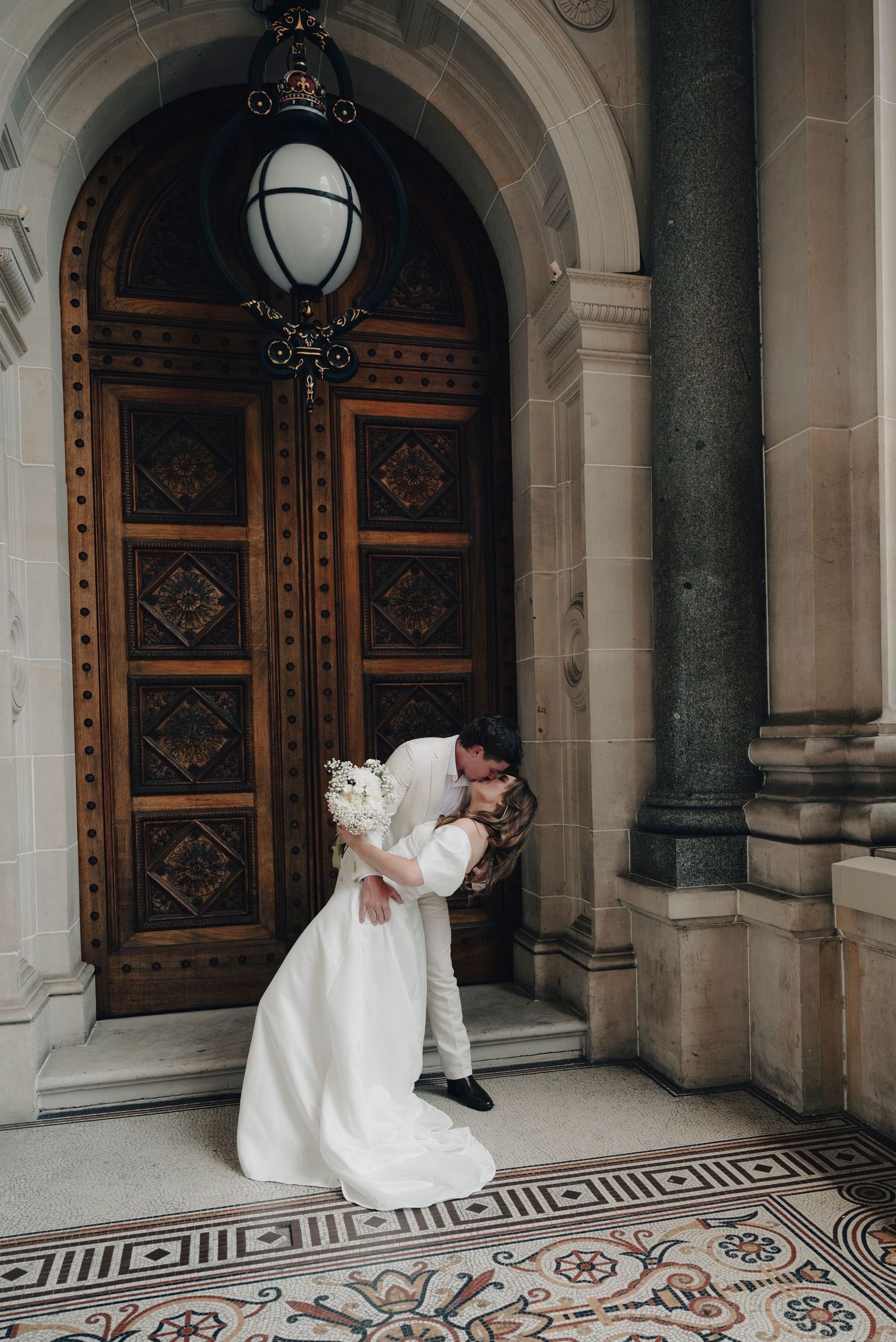 A bride and groom share a romantic moment in an elegant historic building with ornate tiled floors and wooden doors.