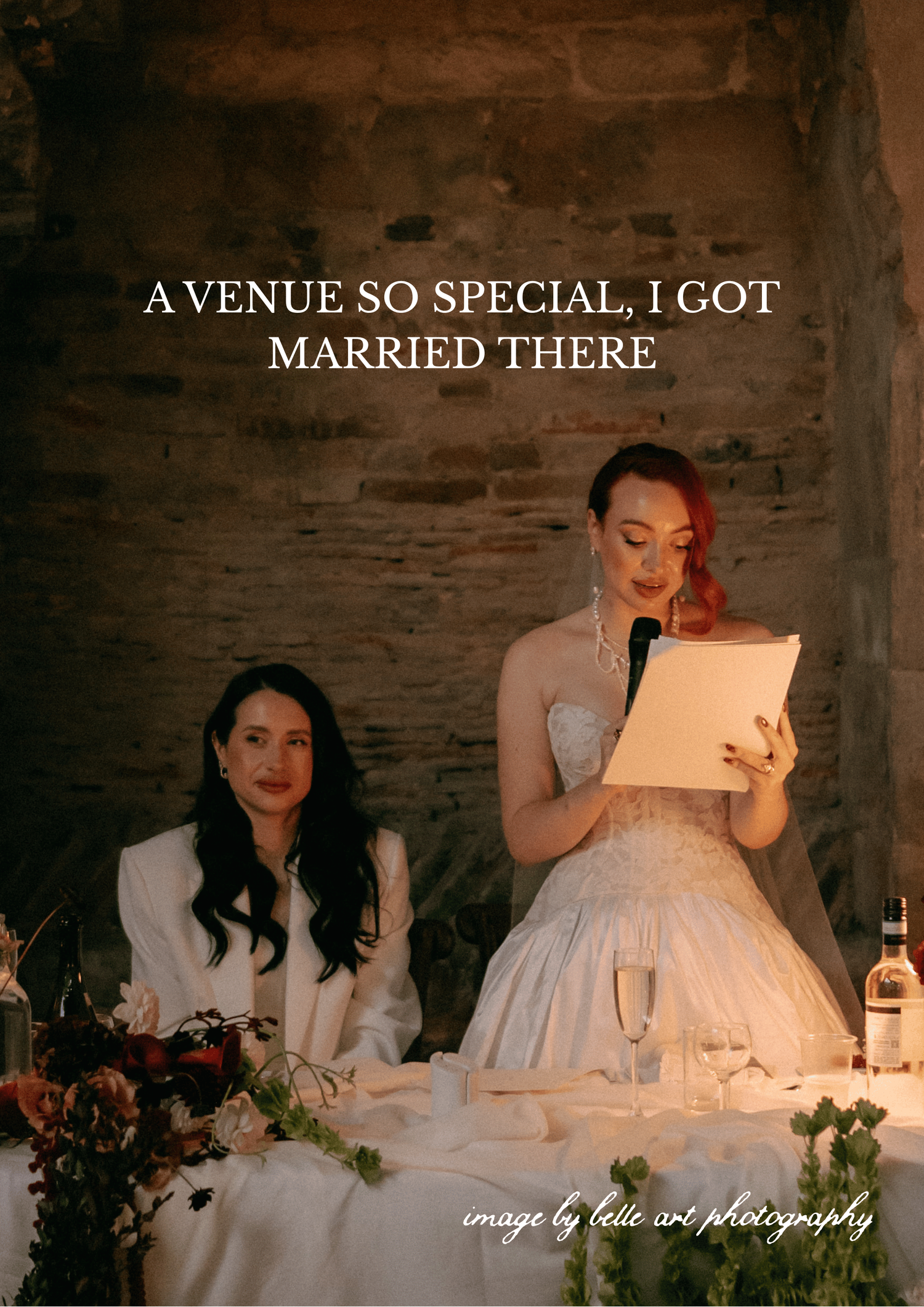 Two people in wedding attire seated at a decorated reception table while one reads from a paper in warm lighting.