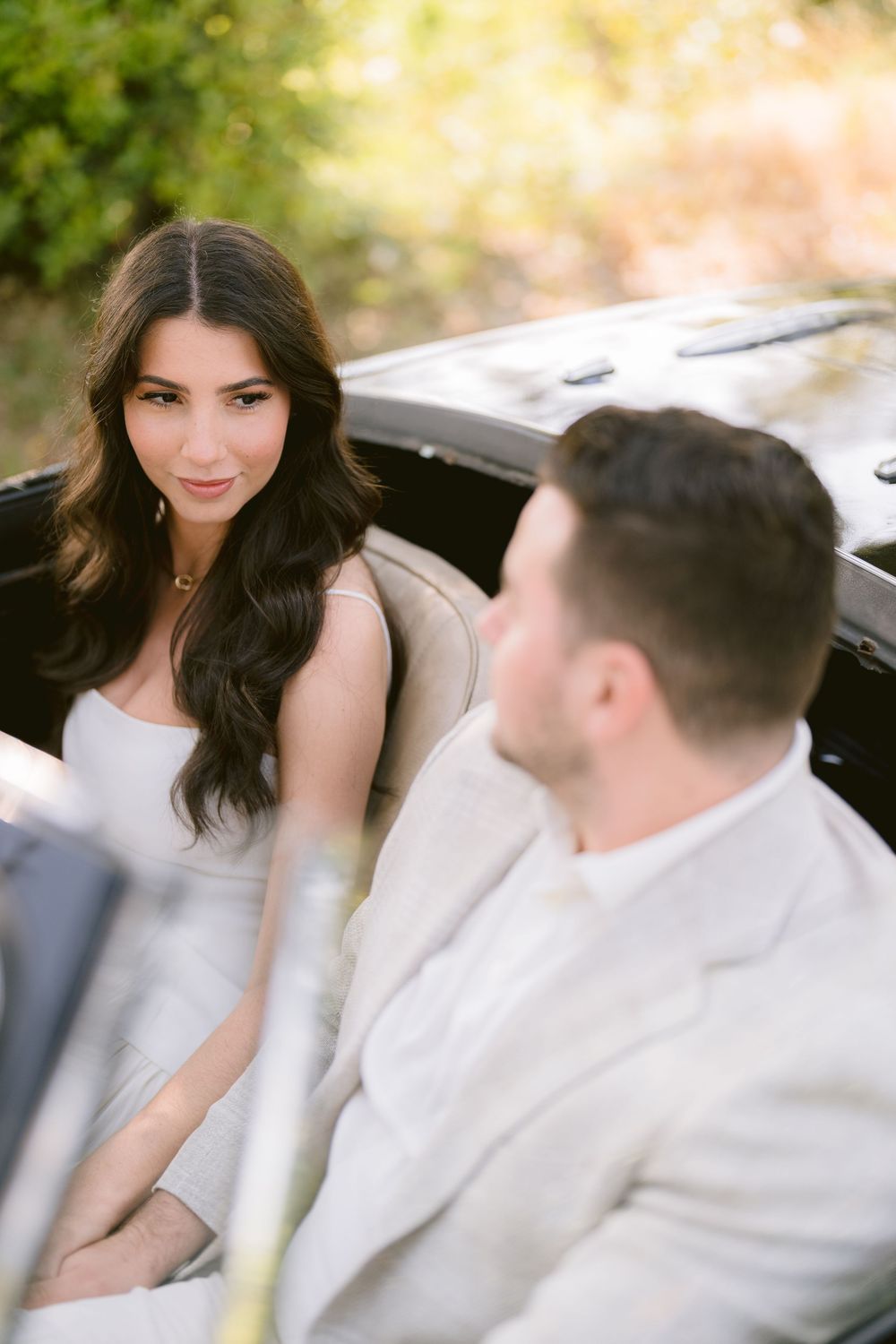 A couple sits together in a vintage car, with the bride wearing a white dress and the groom in a light colored suit.