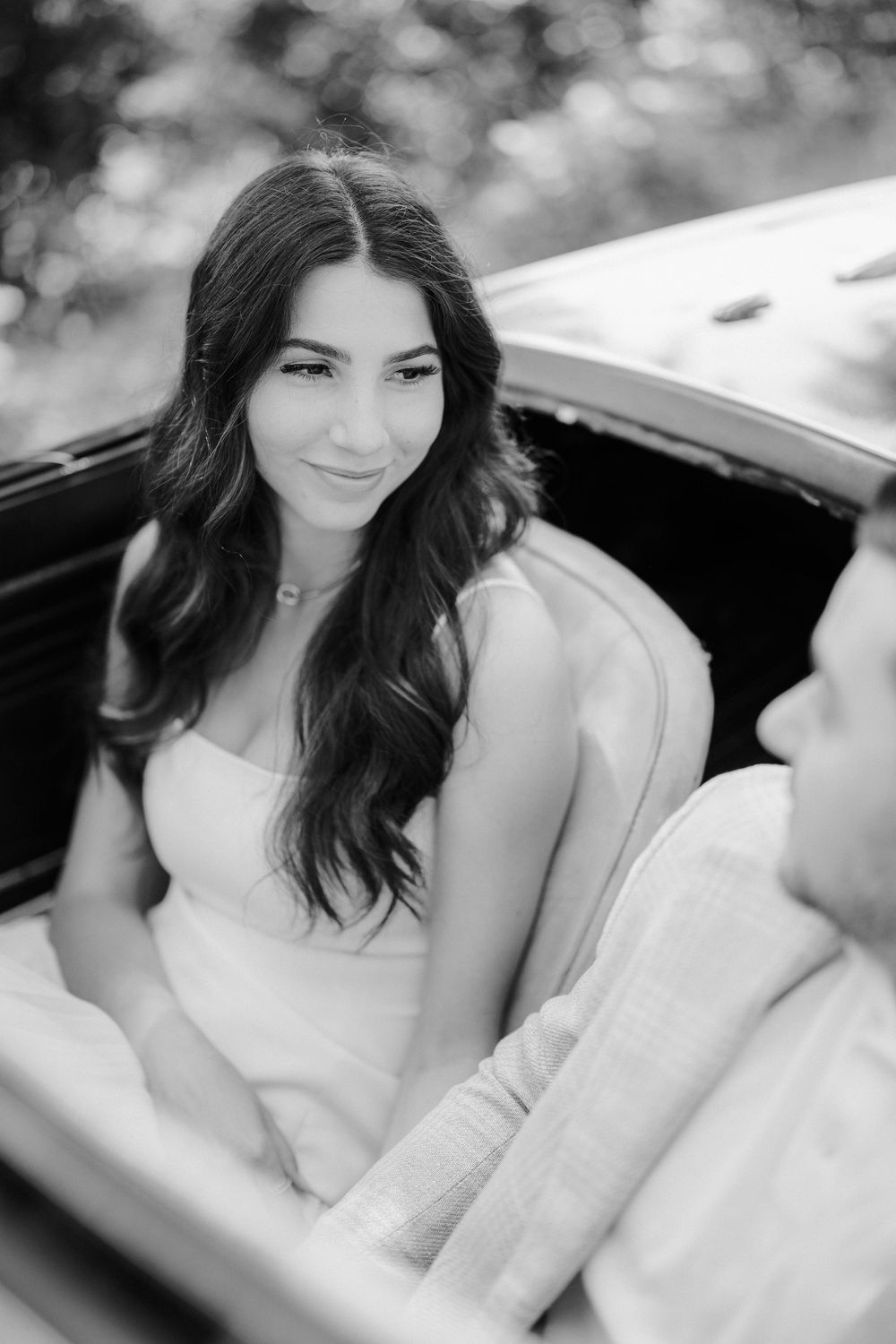 Black and white portrait of a woman with long dark hair sitting in a car wearing a white dress.