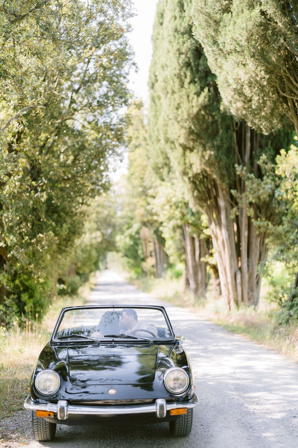 Classic black Jaguar E-Type convertible parked on a tree-lined country road on a sunny summer day.