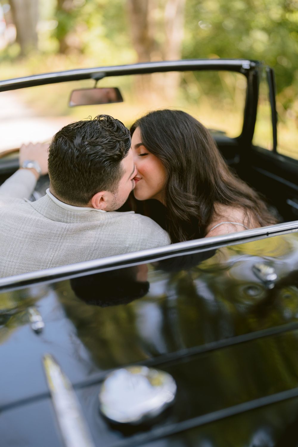 A romantic couple sharing a kiss in a black convertible car on a sunny day.