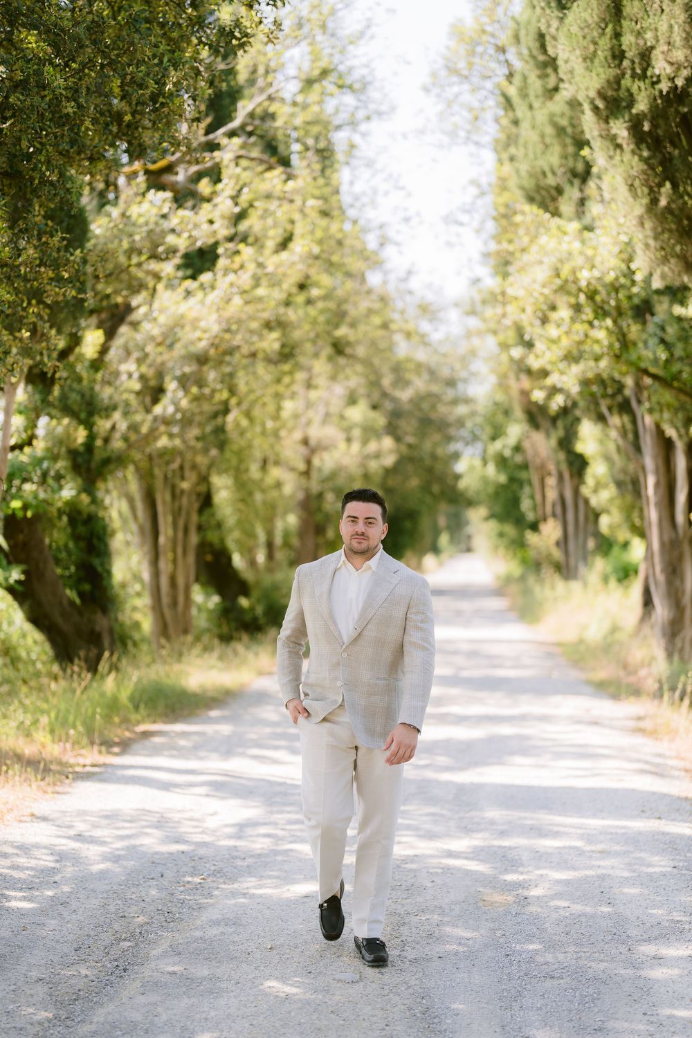 A person in a light gray suit walks along a tree-lined gravel path on a bright sunny day.