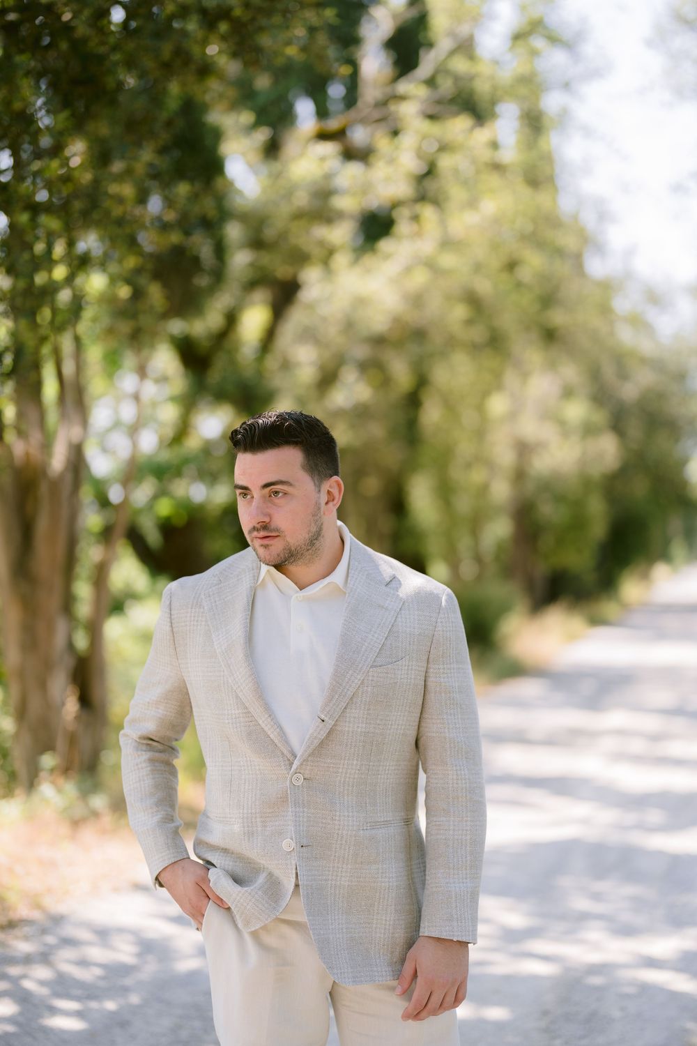 A person wearing a light beige suit poses outdoors on a tree-lined path in natural lighting.