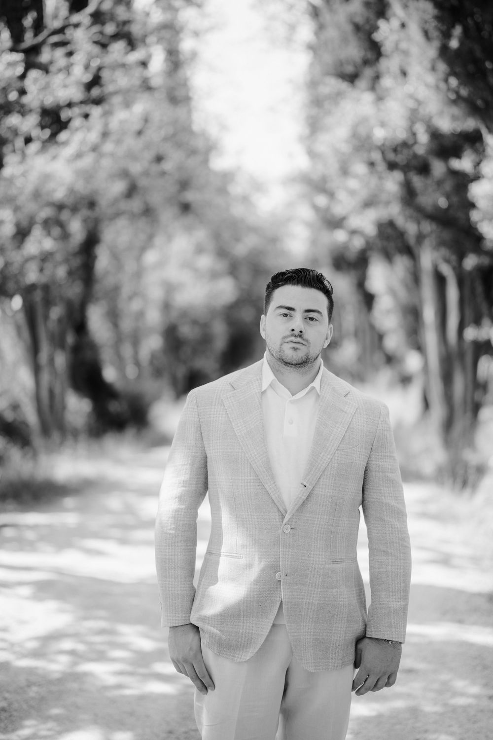 Black and white portrait of a person in a light colored suit jacket standing outdoors in a wooded setting.
