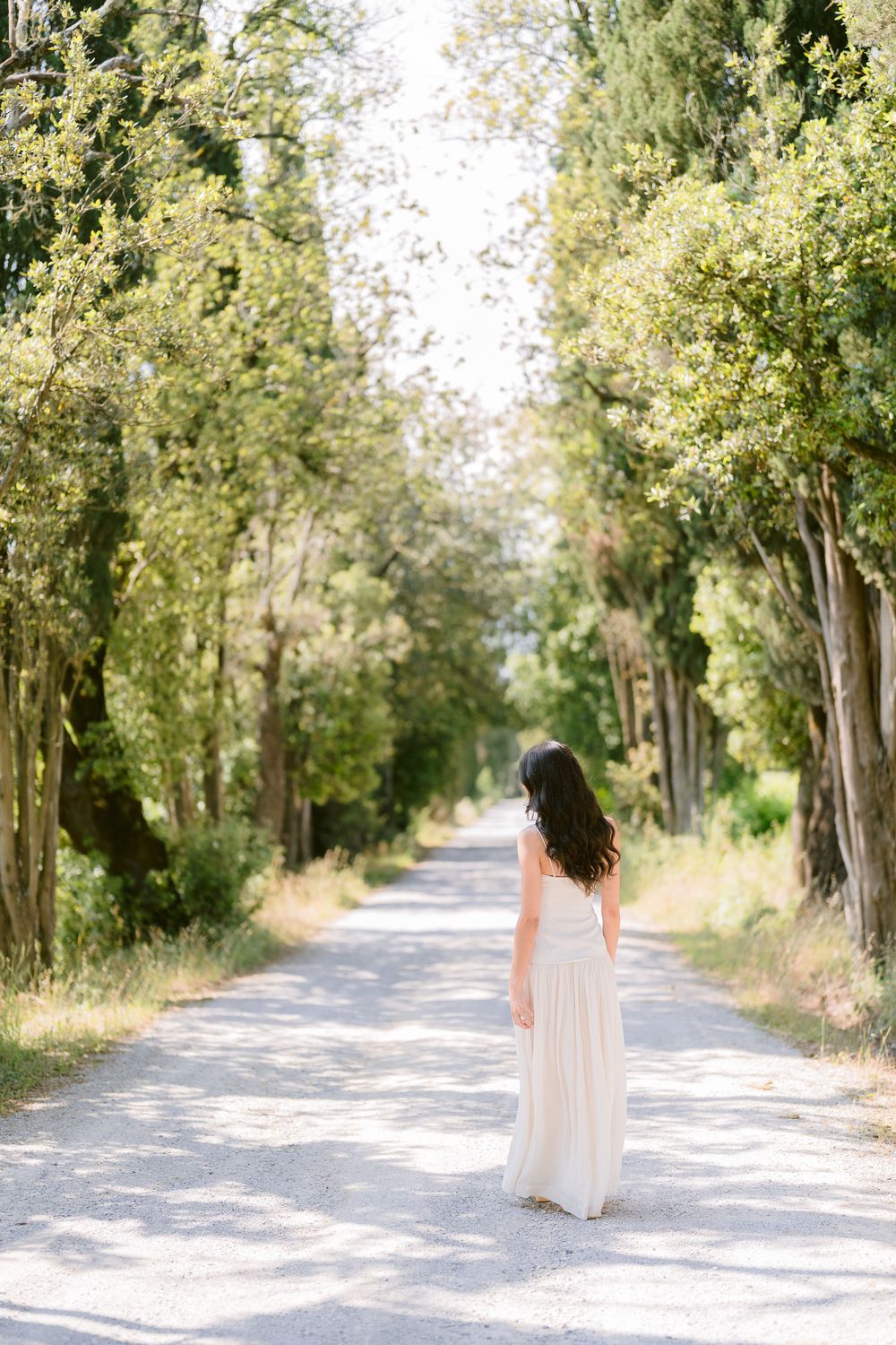 A tree-lined country road with a figure in a white dress walking away into the distance on a bright sunny day.
