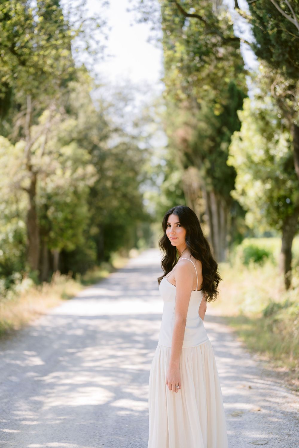 A woman in a flowing white dress stands on a tree-lined dirt path on a bright sunny day.