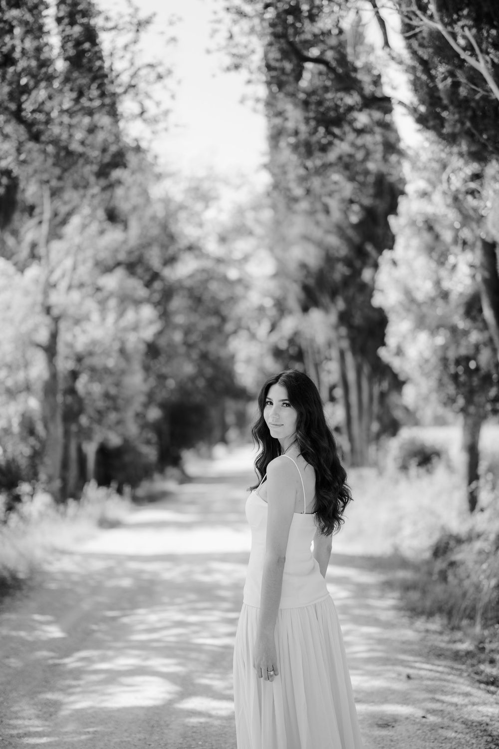 A black and white photograph of a tree-lined path with dappled sunlight creating a dreamy, ethereal atmosphere.