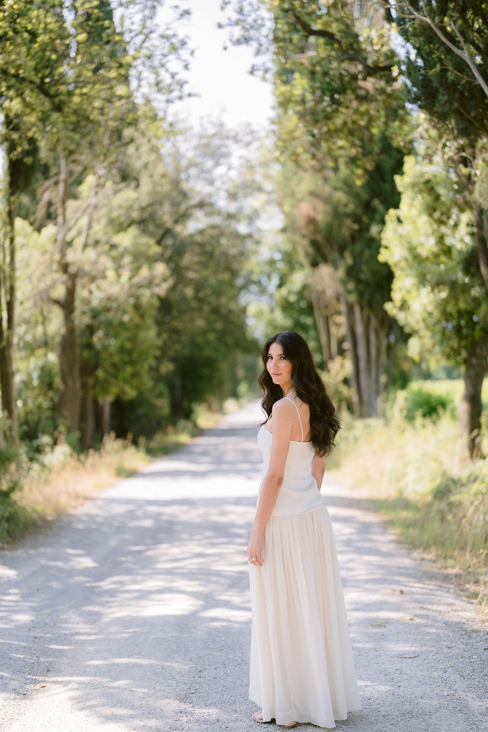 A person in a flowing white dress walks down a tree-lined gravel path on a bright summer day.