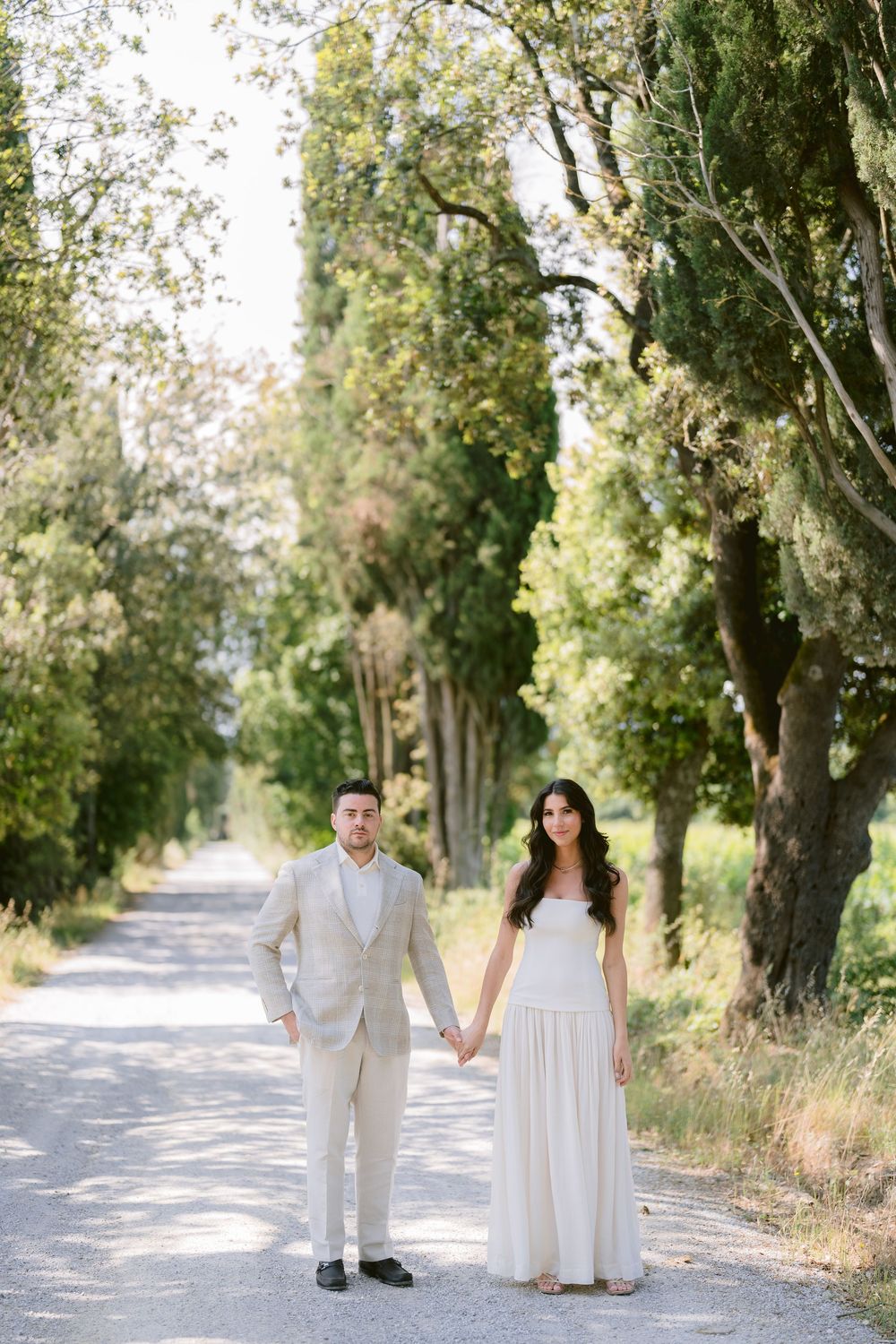 A couple in white outfits walks hand in hand down a tree-lined path on a sunny summer day.