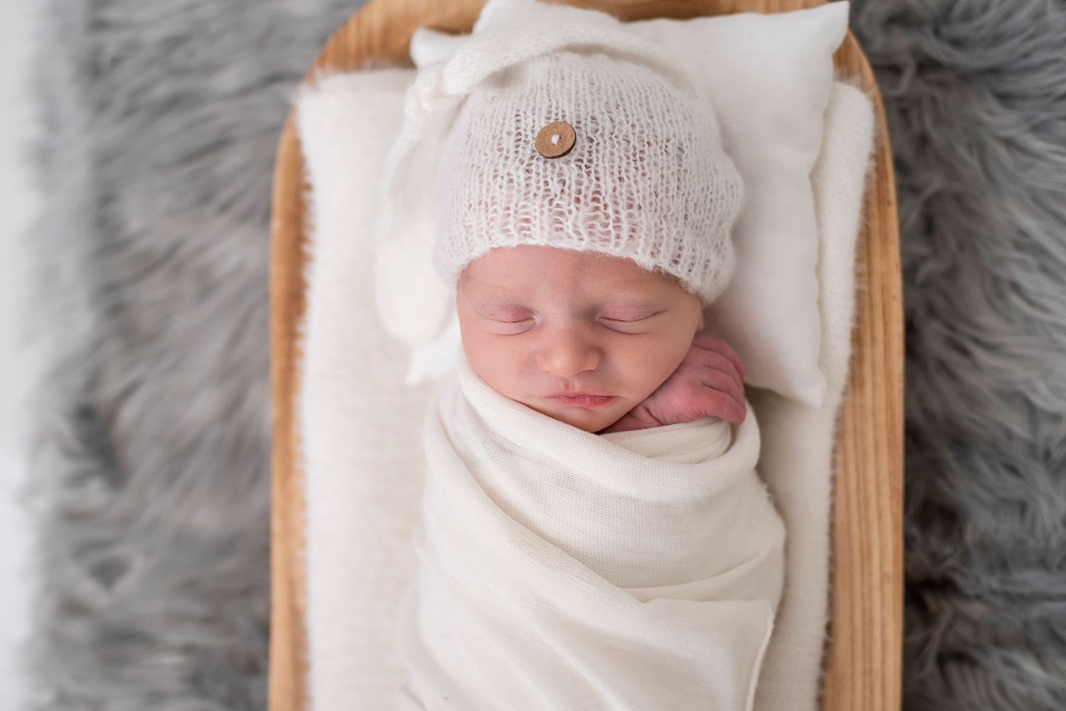 Newborn baby sleeping peacefully in a white knit hat and swaddle blanket against a grey background.