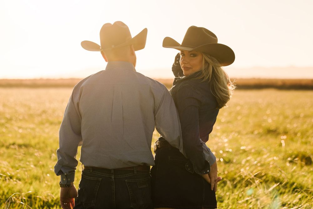 A couple in cowboy hats stands together in a golden field at sunset, viewed from behind.