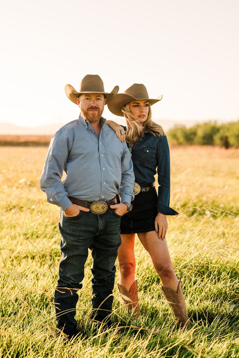 A western couple in cowboy attire poses together in a sunlit field at golden hour.