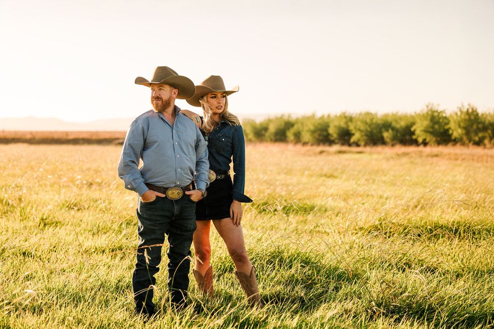 A couple in western attire walks together through a golden wheat field at sunset.