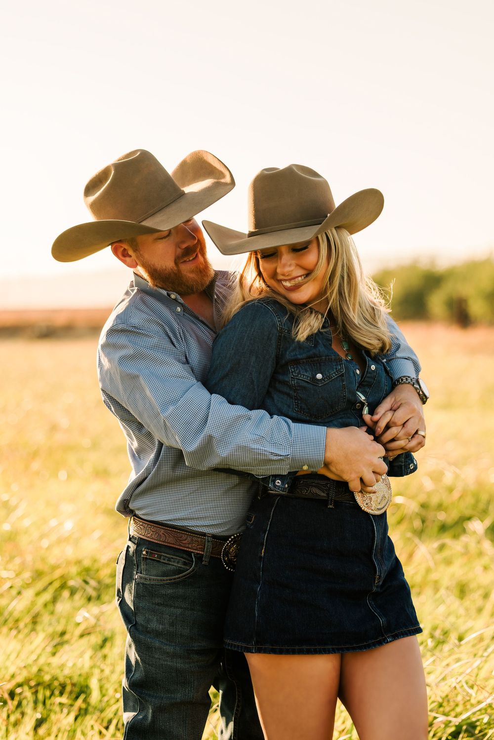 A couple embraces playfully in a sunlit field during golden hour wearing casual western attire.