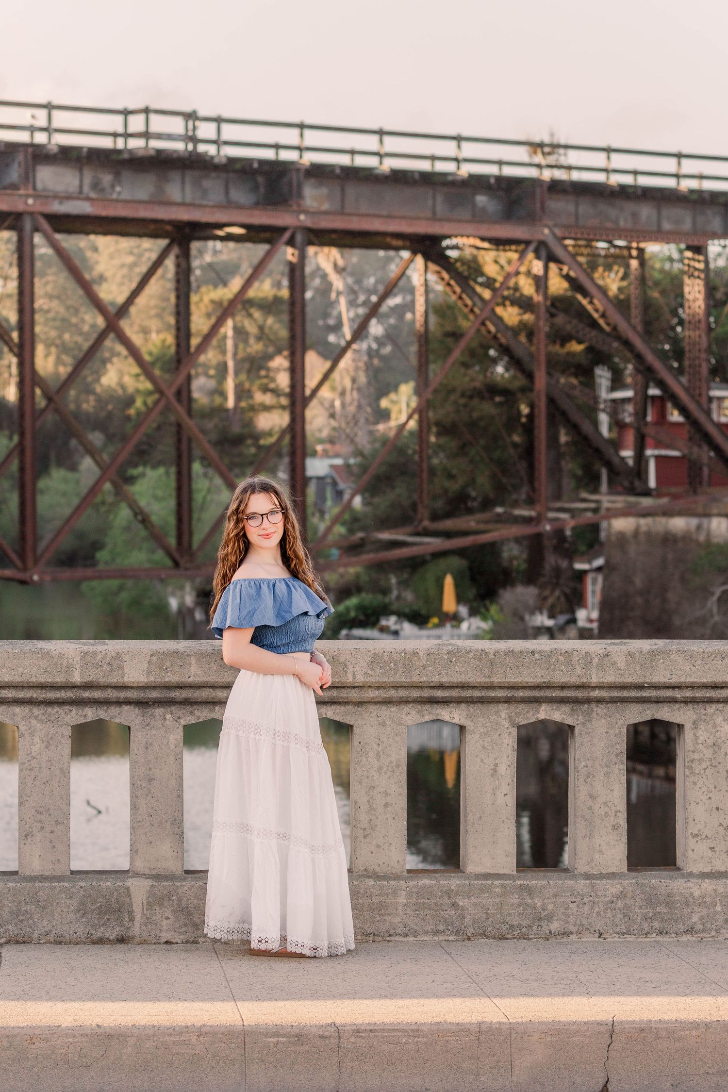 A person in a white tulle skirt and denim jacket poses near a rustic bridge structure at sunset.