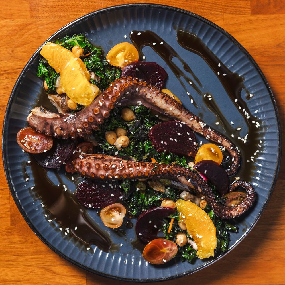 Grilled octopus with roasted potatoes and herbs served on dark blue plates against a wooden surface.