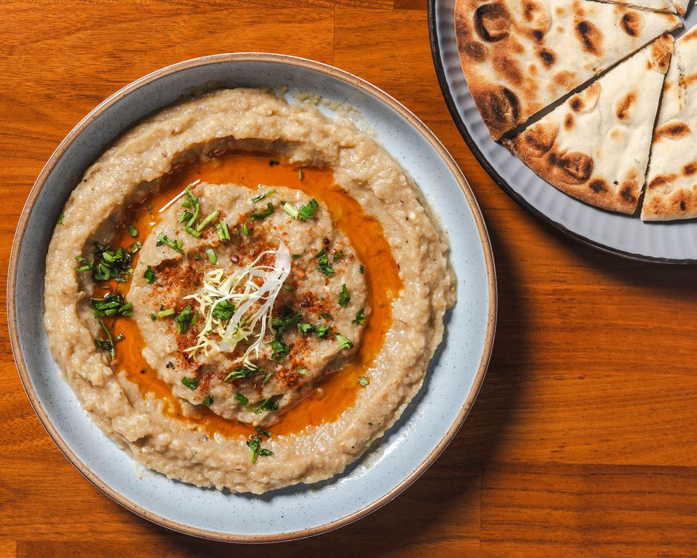 A row of creamy hummus bowls garnished with olive oil, spices and herbs on a wooden table surface.