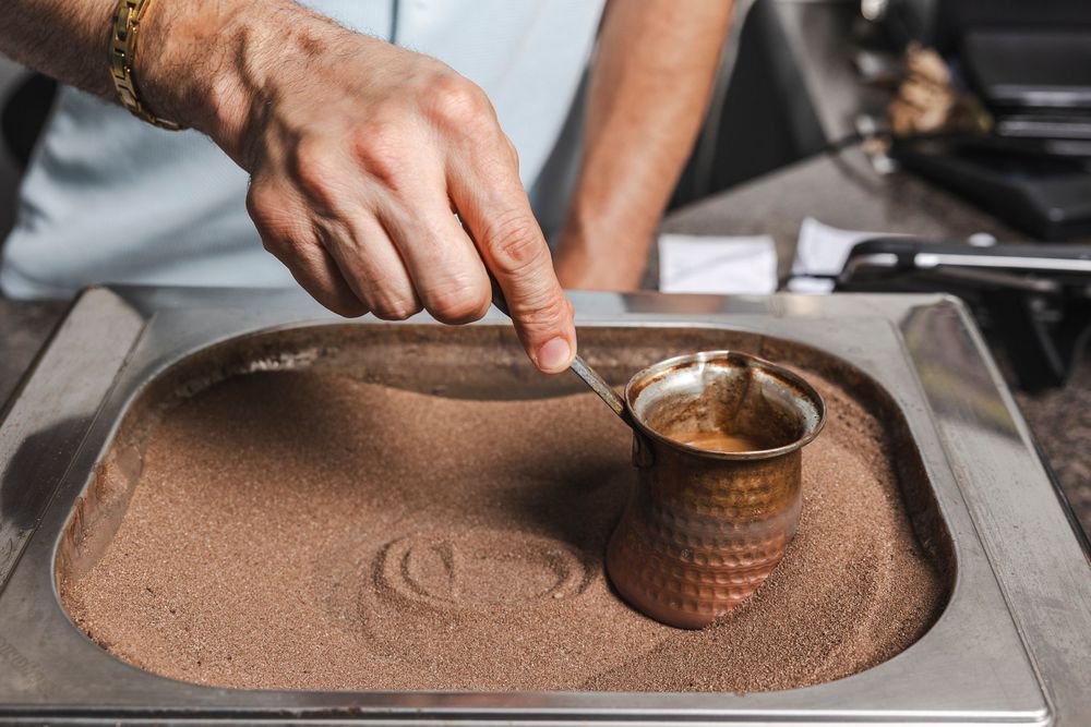 Hand preparing traditional Turkish coffee in hot sand, with copper cezve pot nestled in brown sand on metal tray.