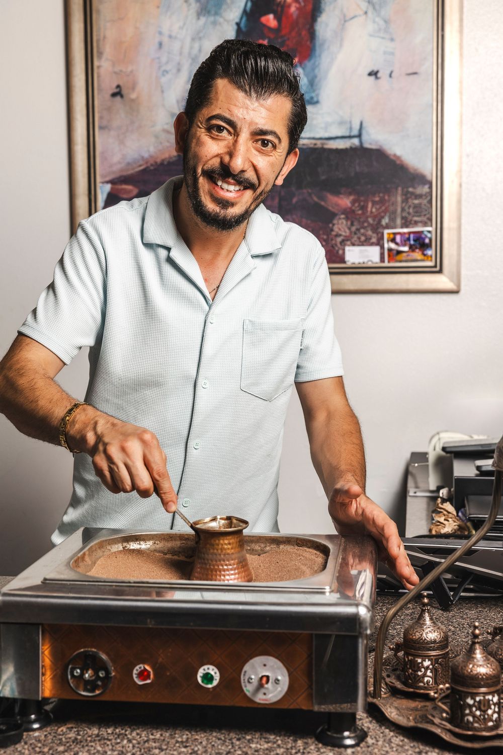 A person in a white polo shirt prepares traditional Turkish coffee using a copper cezve pot on a stovetop.