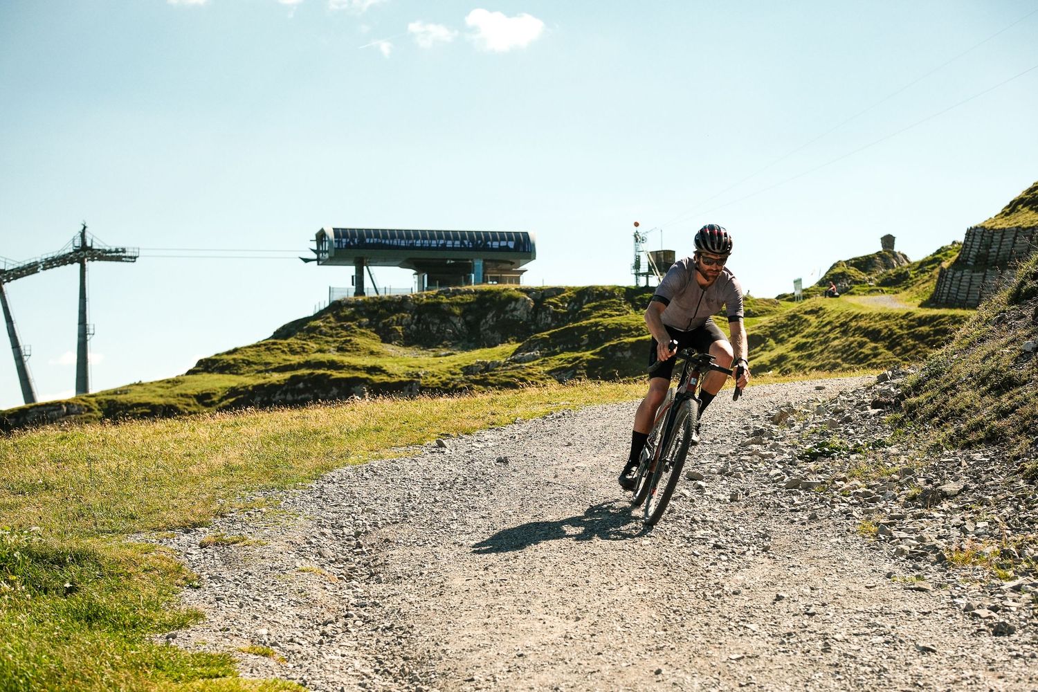 Mountain bikers navigate a steep, winding dirt trail through scenic alpine terrain on a sunny day.