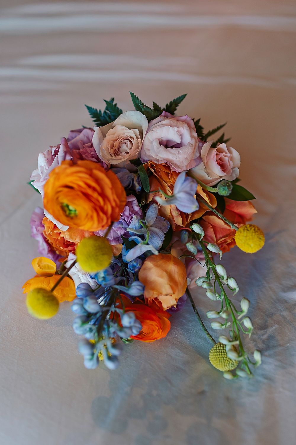 A vibrant floral arrangement featuring orange ranunculus, pink roses, yellow craspedia, and delicate blue blooms on a gray surface.