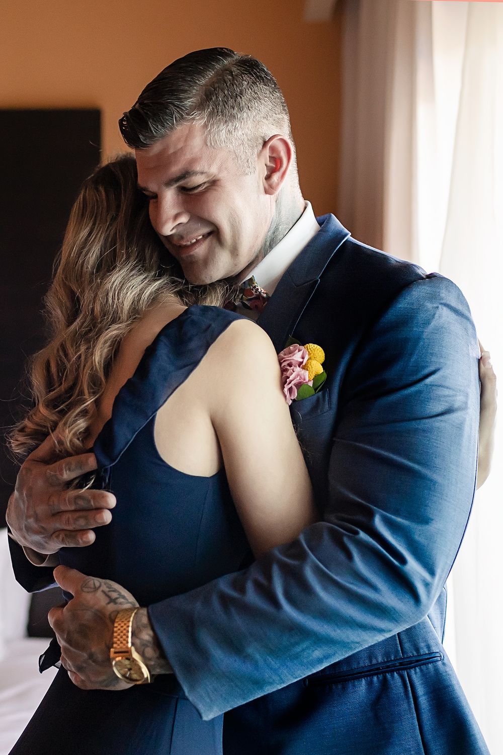 A tender embrace between two people in formal attire, with the person wearing a navy blue suit and floral boutonniere.