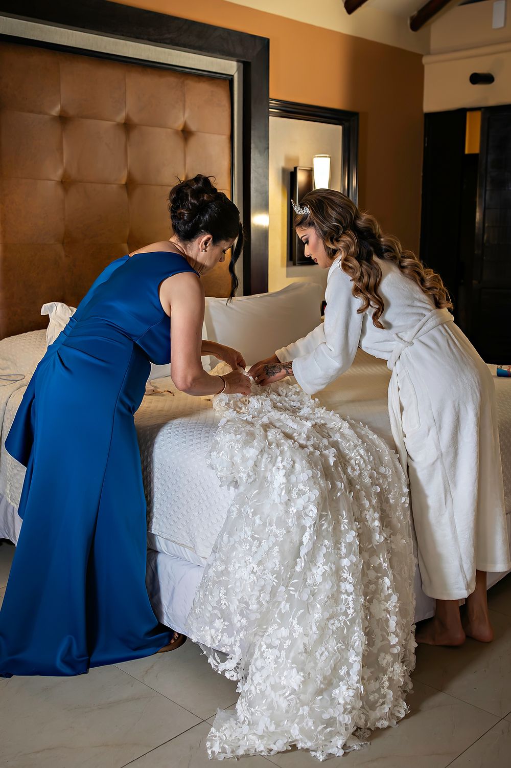 Two people in blue dresses helping arrange a lace wedding gown on a bed.