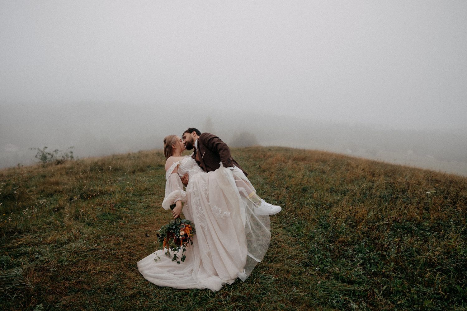 A couple in wedding attire embraces on a misty hillside surrounded by wildflowers at sunset.