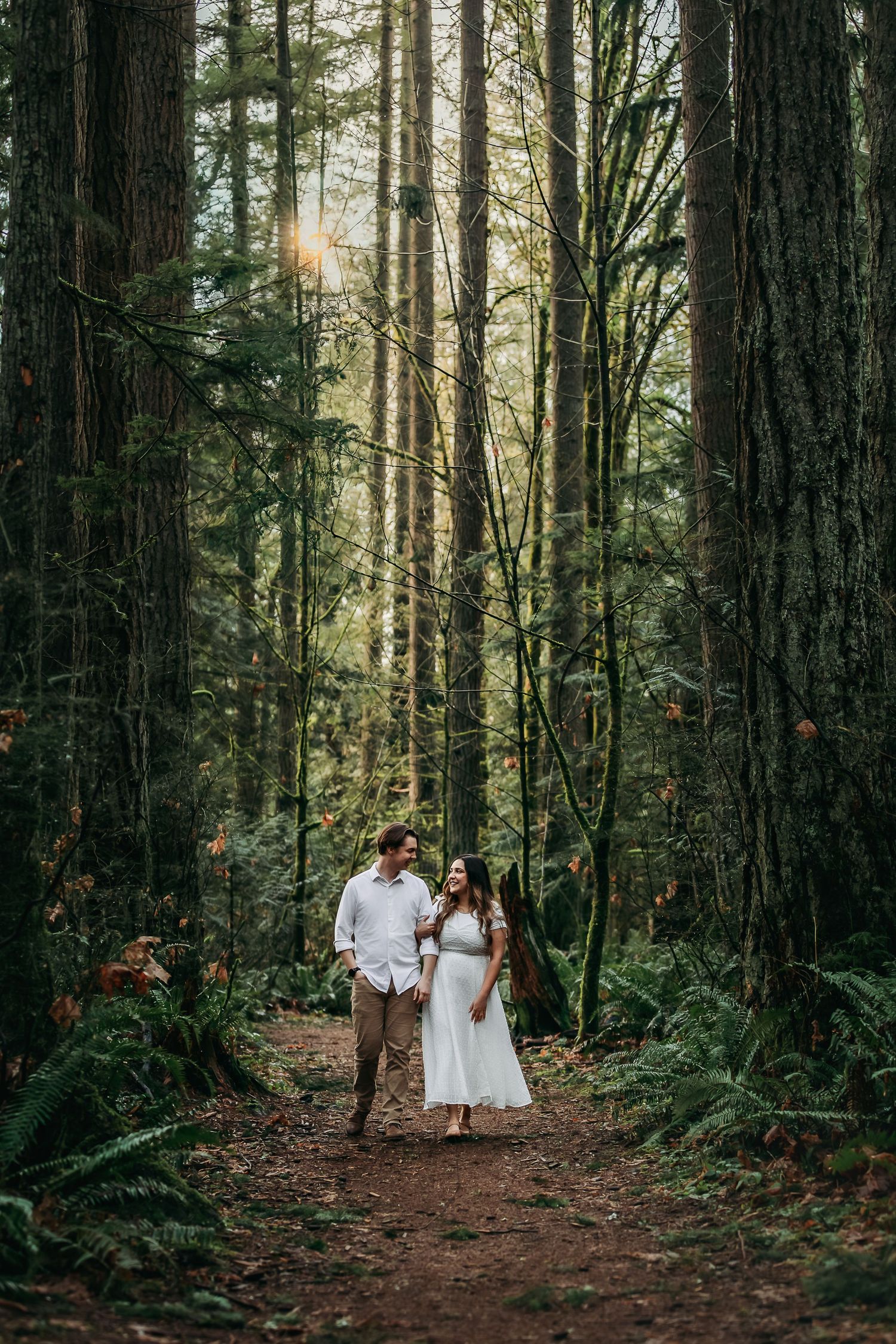 Couple walking through towering evergreen forest on dirt path with filtered sunlight.