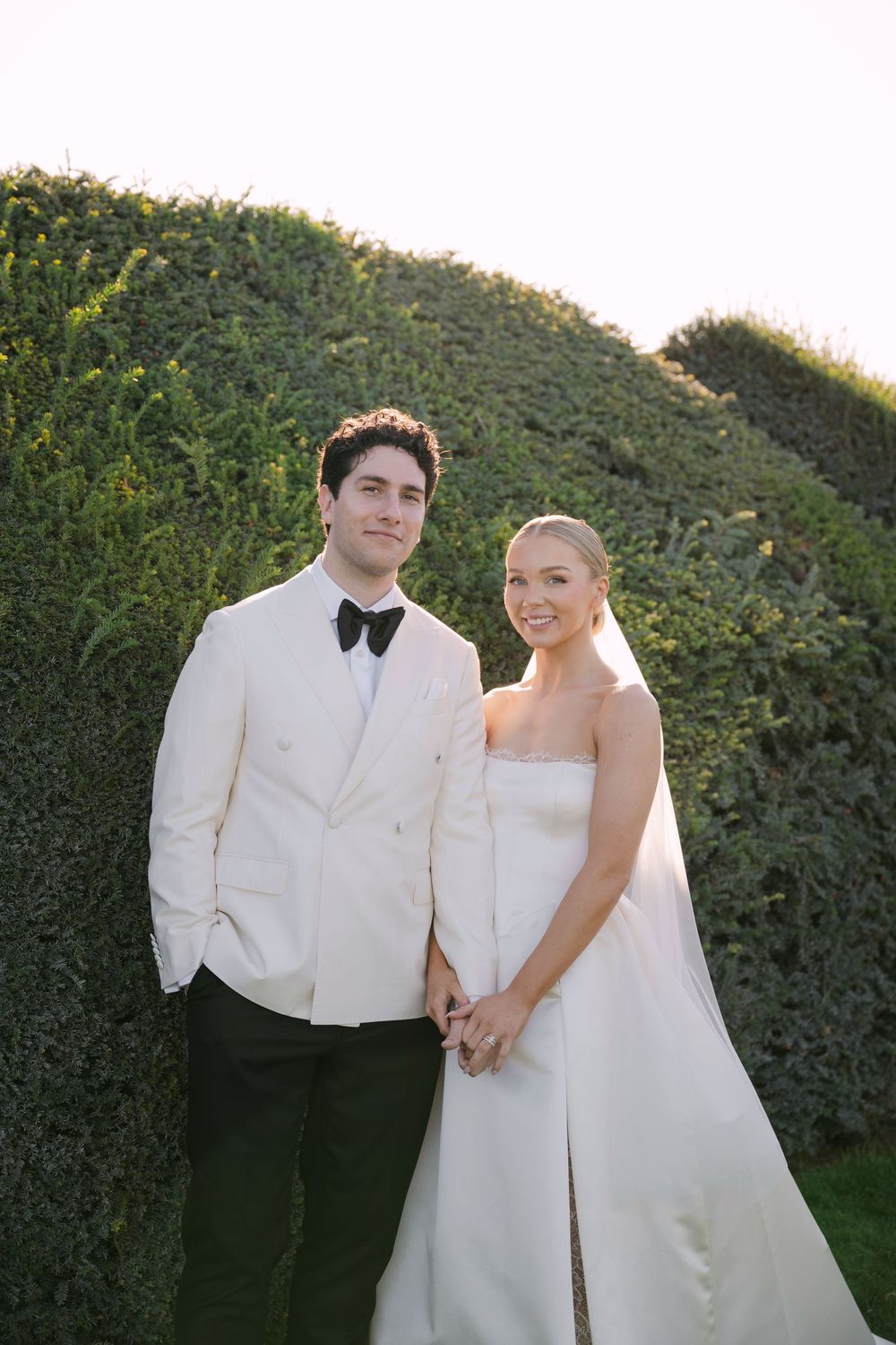 A couple in formal white wedding attire poses together in front of a green hedge on their special day.