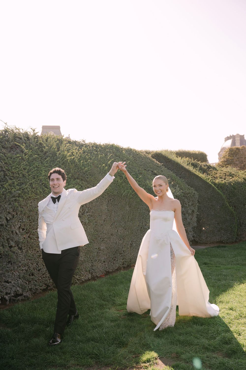 A bride and groom playfully pose and dance together on their wedding day in a garden setting with hedges.