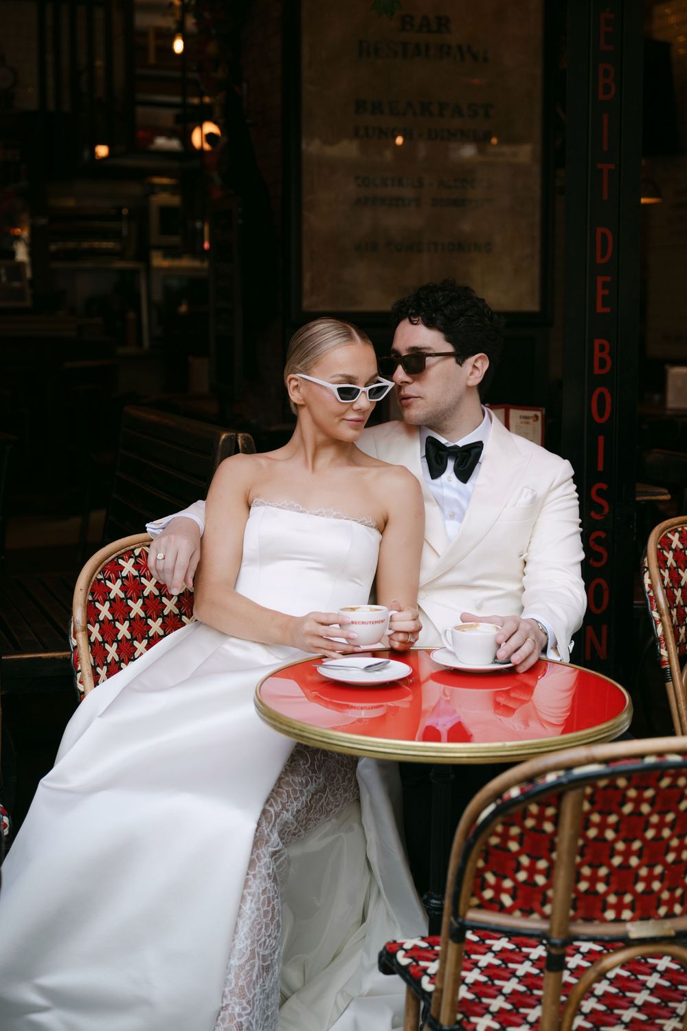 A couple in wedding attire shares an intimate moment at a Parisian cafe with red chairs and a small table.