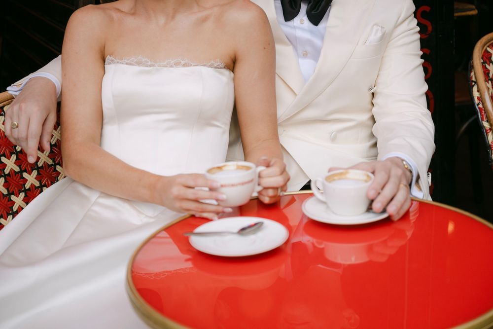 Hands holding white teacups and saucers against a bright red table surface in a cafe setting.