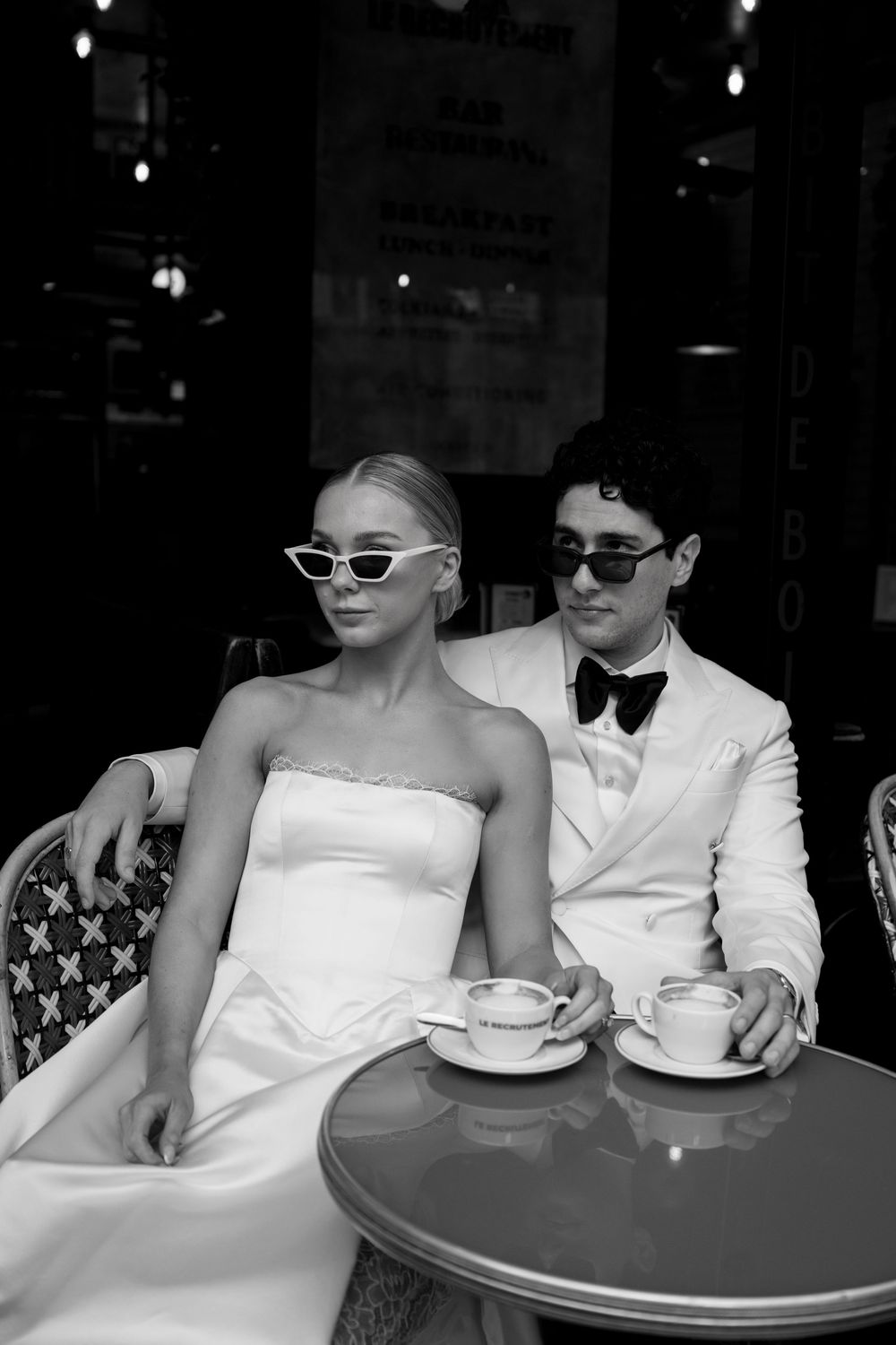 A black and white photo shows two people in formal white attire sitting at a cafe table with coffee cups.