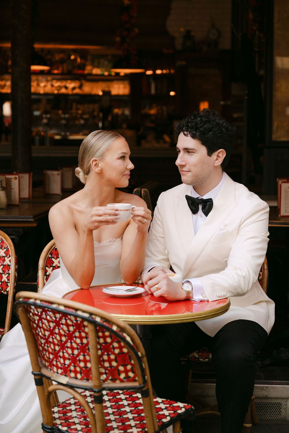 A couple sits at a romantic outdoor cafe with red bistro chairs sharing an intimate moment in Paris.