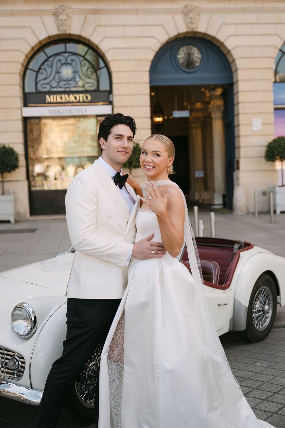 A romantic wedding couple poses near a vintage white car outside an elegant building with classic architecture.