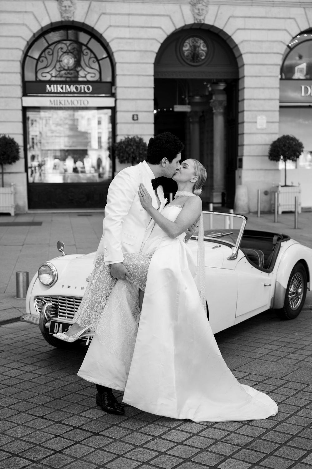 A romantic black and white wedding photo with a vintage white convertible car parked outside a Mikimoto storefront.