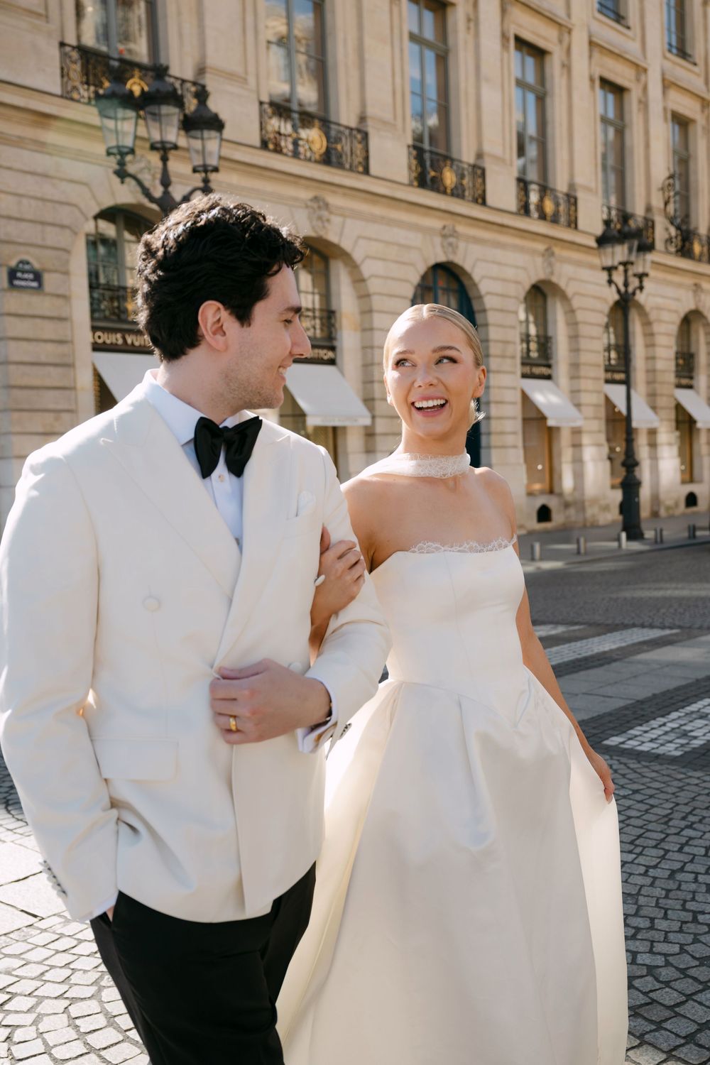Joyful wedding couple in white attire share candid laughing moments on a cobblestone street in Paris.