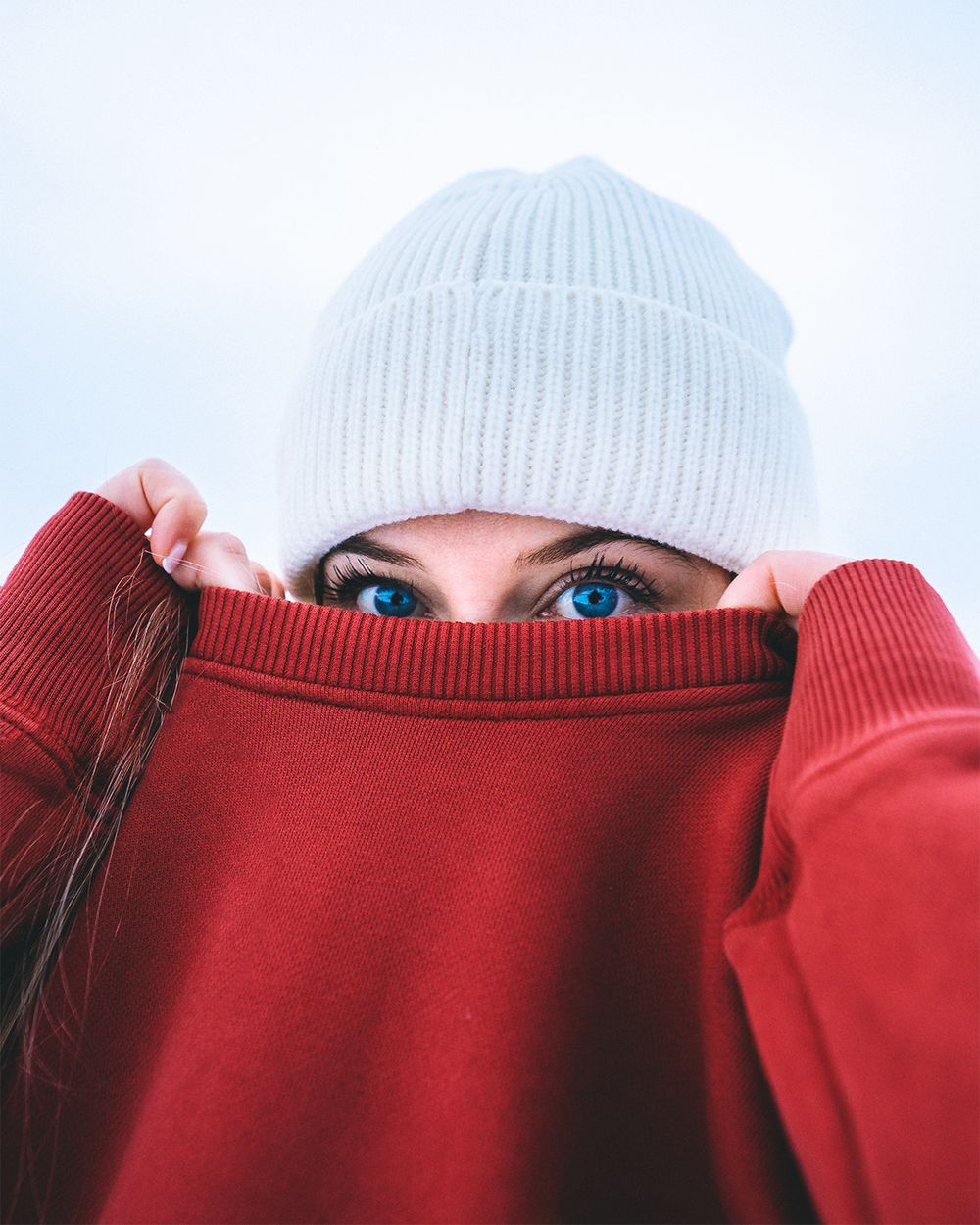 A person wearing a red sweater and white beanie pulls the collar up to reveal just their bright blue eyes.