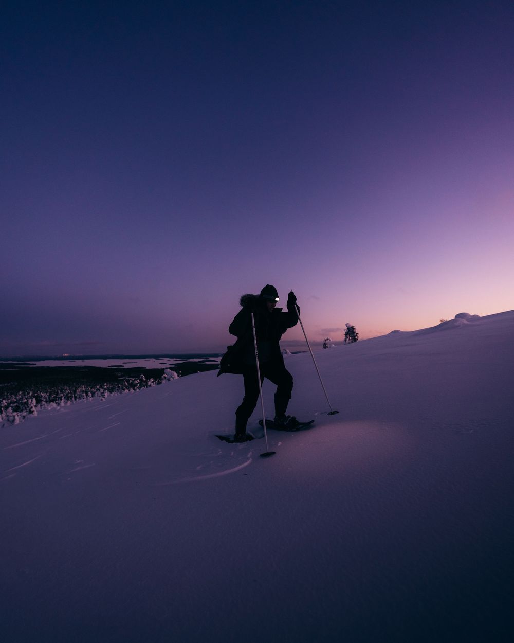 Silhouetted skiers traverse a snowy slope against a dramatic purple twilight sky in winter.