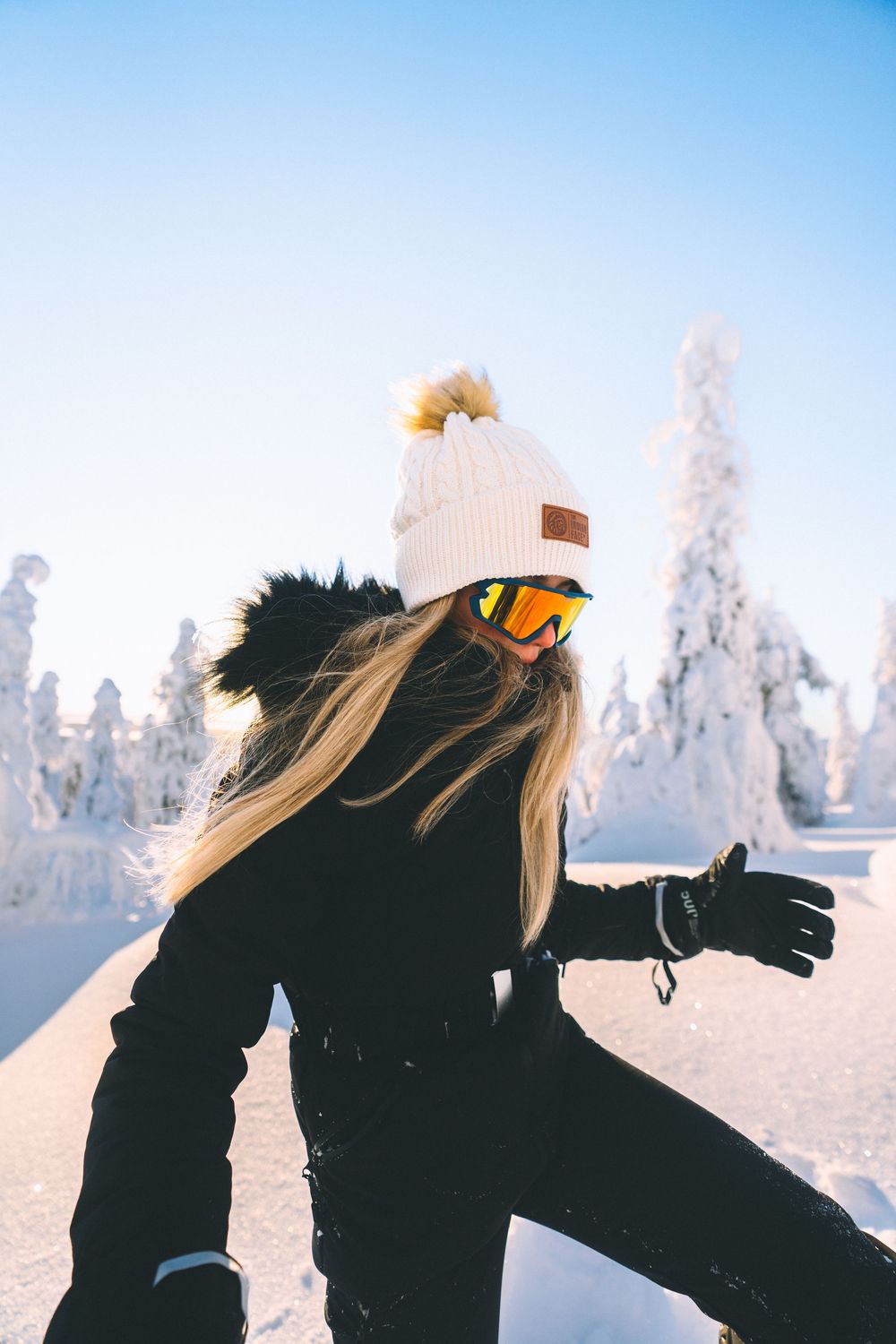 Person in black winter clothing and beanie throws snow into the air against a snowy winter landscape at sunset.