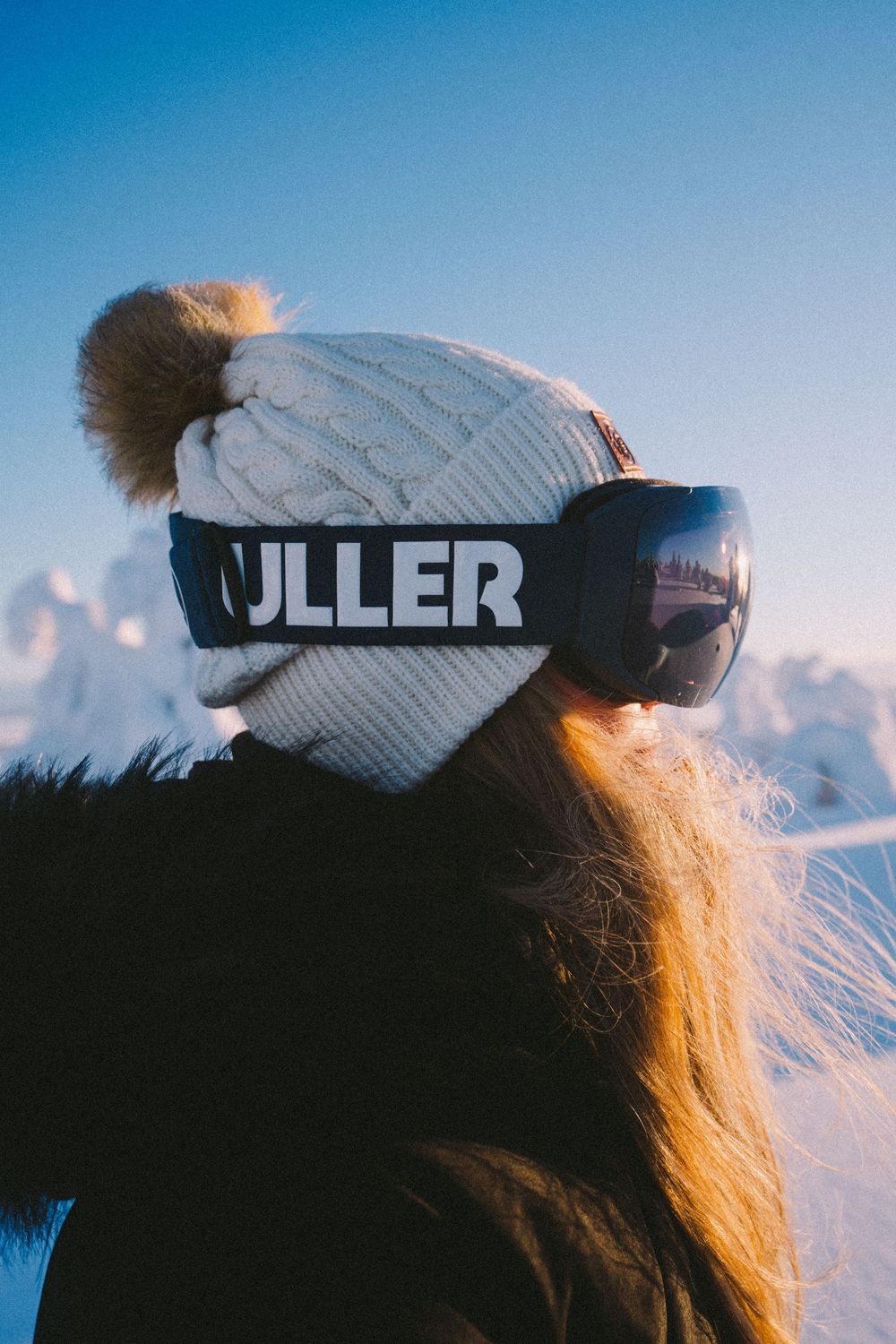 Close-up view of someone wearing a white knit beanie and ski goggles against a bright blue winter sky.