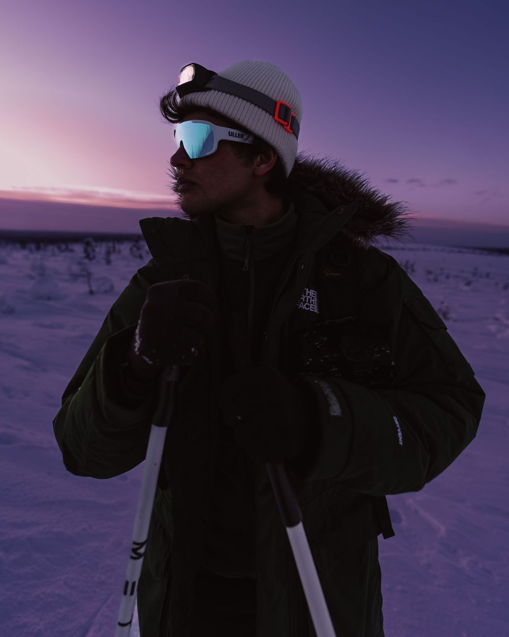 Winter adventurer in black puffer jacket and beanie stands against snowy landscape during purple sunset.