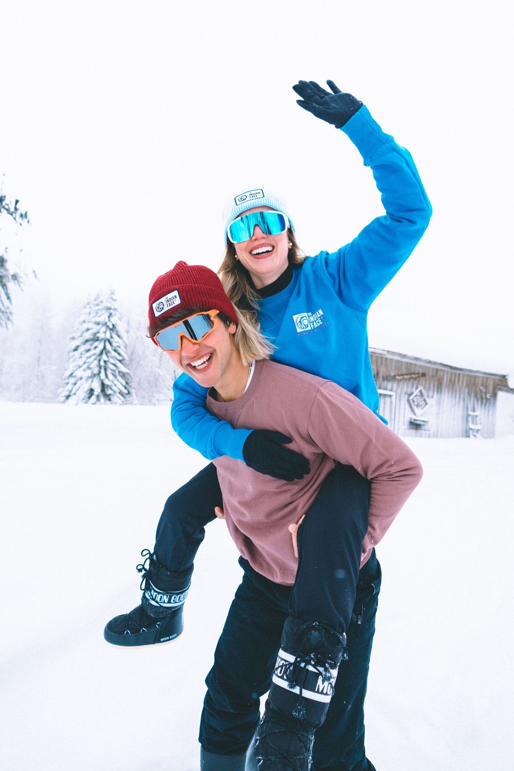 Two snowboarders in winter gear laugh and pose together on snowy slopes during a bright winter day.