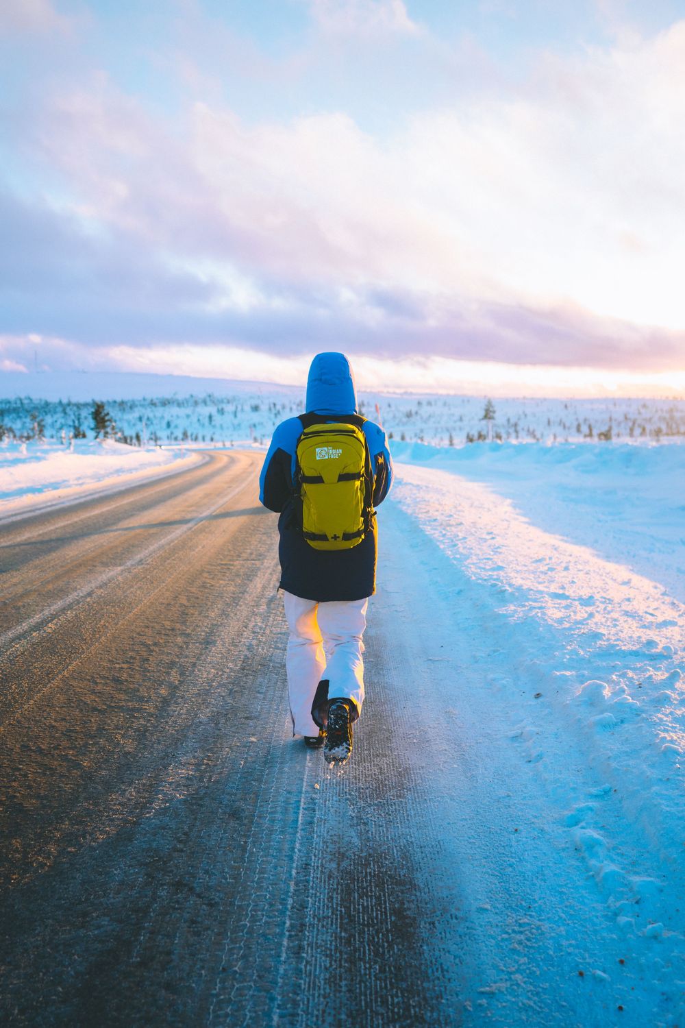 A person with a yellow backpack walks on a snowy road during sunset in a winter landscape.