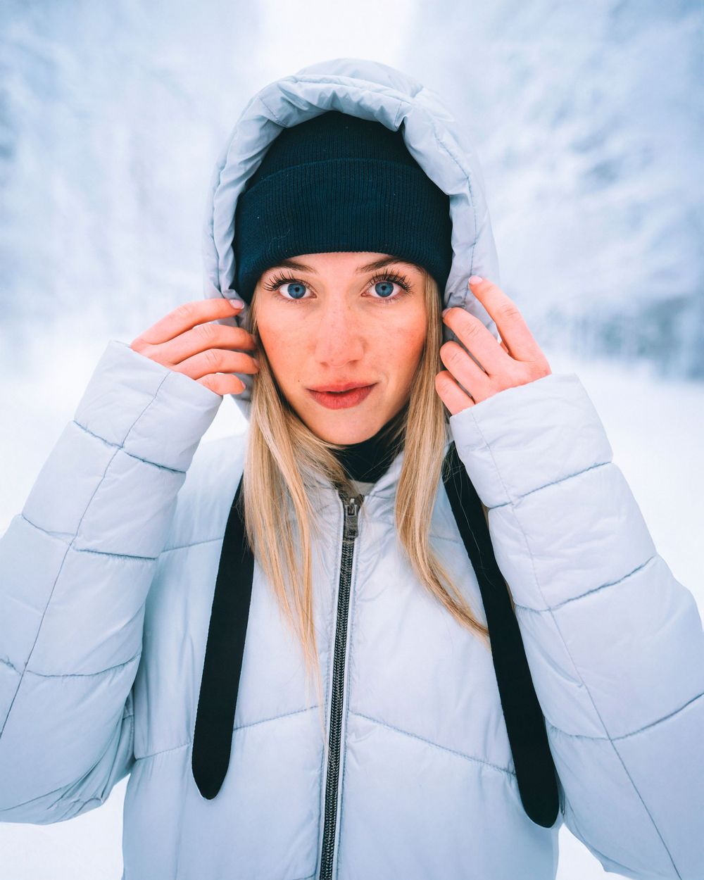 Person in white winter puffer coat and black beanie poses playfully in snowy outdoor setting.